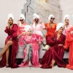 Eight women stand wearing stain gowns with towels wrapped on their heads and shades on